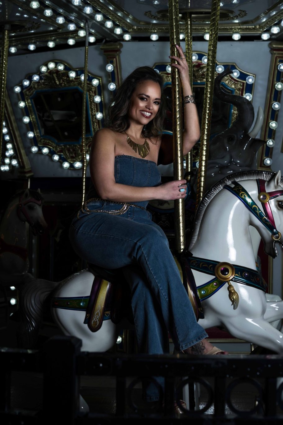 Woman in a denim jumpsuit riding a white carousel horse at night.