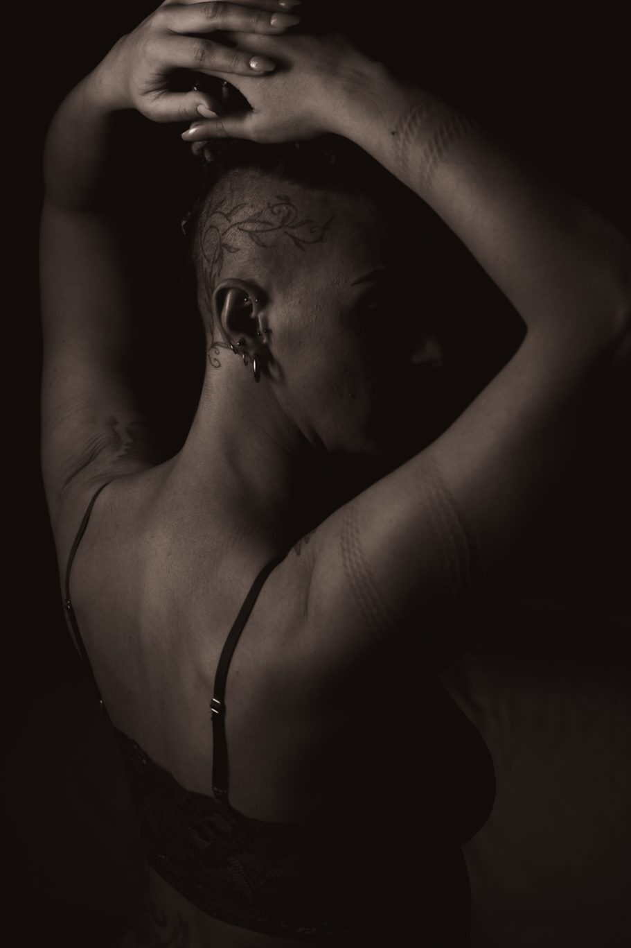 A person with short hair, seen from behind, poses with arms raised in a dark setting.