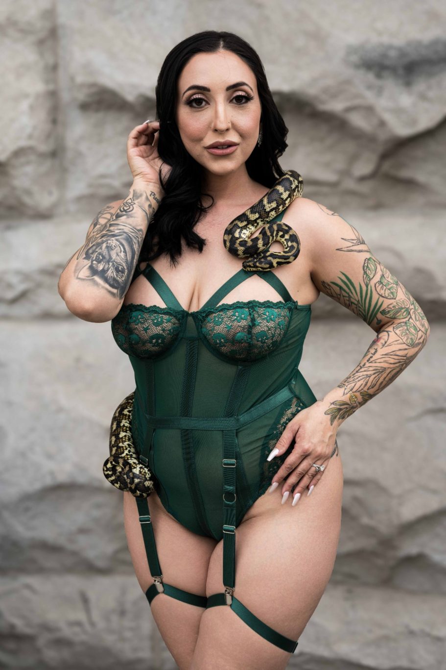 Woman in a green lingerie outfit with snakes draped around her, standing against a stone wall.
