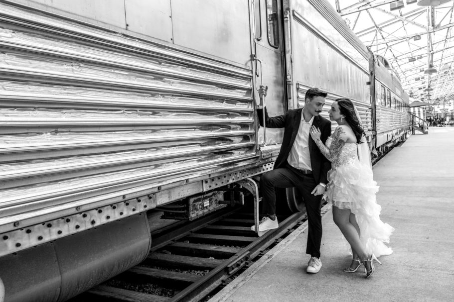 A couple in formal attire poses playfully by a vintage train in a industrial setting.