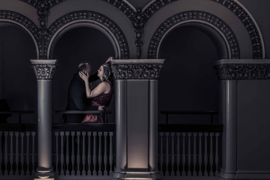 A couple embracing on a balcony with ornate arches and dramatic lighting.