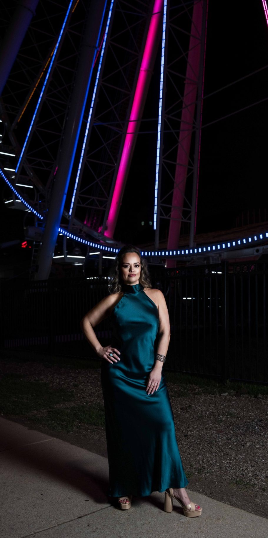 Woman in a teal dress poses beside a brightly lit Ferris wheel at night.