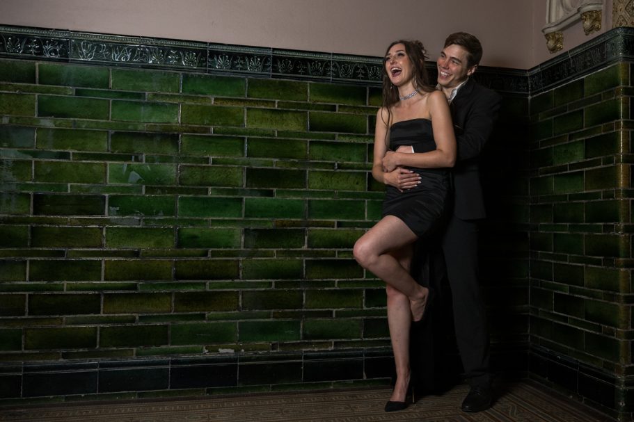 A couple poses against a green tiled wall, with the woman in a black dress and the man in a suit.