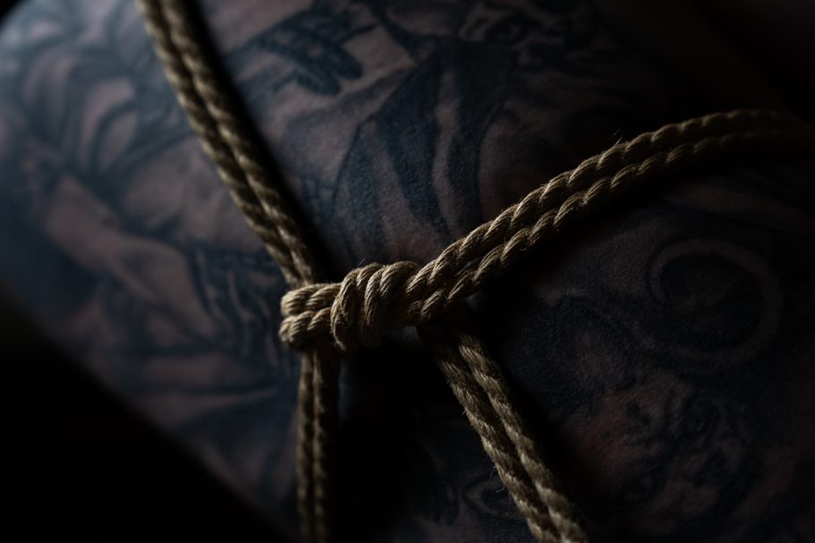 A close-up of a tattooed arm with a knot of brown rope tied around it.