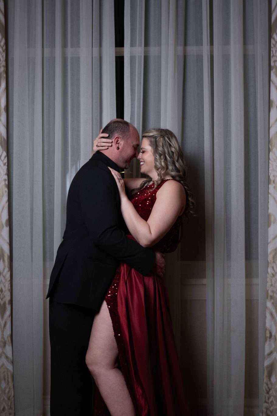 Couple embracing, dressed elegantly, with soft lighting and curtains in the background.