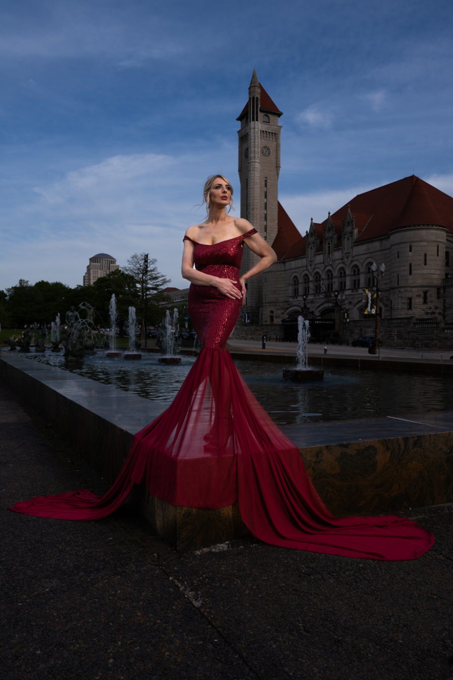 A model in a flowing red gown poses by a fountain with a historic building in the background.